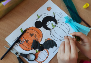 Dzieci wykonują prace plastyczne przy użyciu czarnego kartonu - kot, pomarańczowej bibuły - dynia, białej bibuły – duszek, czarnej bibuły – nietoperz, czarnego kartonu – pająki, zielonej kartki - listki.