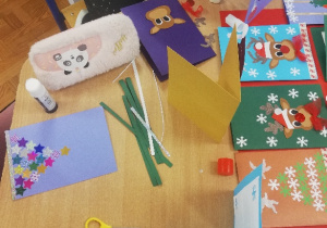 Dzieci wykonują świąteczne kartki z kartonów i kolorowych kartek, ozdabiają je świecącymi gwiazdkami i śnieżynkami. Powstają wesołe renifery, bałwanki, mikołaje, aniołki, bombki. Dzieci piszą własnoręcznie świąteczne życzenia.