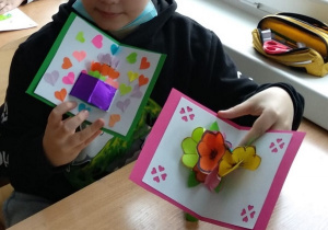 Dzieci prezentują laurki wykonane z kartonu, kwiaty wykonane z kolorowych kartek ksero. Pojedyncze kwiaty sklejone w bukiet. Druga laurka wykonana jest z kartonu z ponaklejanymi kolorowymi serduszkami.