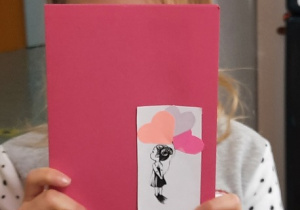 Dziewczynka prezentuje przód laurek. Wydrukowana postać dziecka trzymająca kolorowe baloniki w kształcie serduszek wycięte z kolorowych kartek ksero.