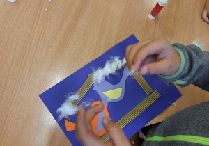 Dzieci podczas zajęć plastycznych w świetlicy wykonują karmniki. Na chabrowej kartce ksero naklejają karmnik wykonany z pofalowanej tektury w karmnikach kolorowe ptaszki powycinane z kolorowych kartek ksero.