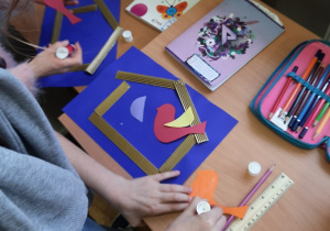 Dzieci podczas zajęć plastycznych w świetlicy wykonują karmniki. Na chabrowej kartce ksero naklejają karmnik wykonany z pofalowanej tektury w karmnikach kolorowe ptaszki powycinane z kolorowych kartek ksero.