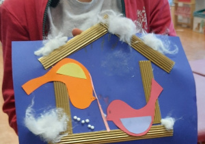 Dzieci prezentują swoje prace plastyczne. Na chabrowej kartce ksero naklejony karmnik wykonany z pofalowanej tektury w karmnikach kolorowe ptaszki powycinane z kolorowych kartek ksero, na daszku karmnika naklejona wata imitująca śnieg.