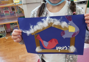 Dzieci prezentują swoje prace plastyczne. Na chabrowej kartce ksero naklejony karmnik wykonany z pofalowanej tektury w karmnikach kolorowe ptaszki powycinane z kolorowych kartek ksero, na daszku karmnika naklejona wata imitująca śnieg.