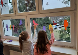 Dzieci pomagają w dekorowaniu świetlicy. Na oknach naklejają kolorowe ptaki wycięte z kartonów.