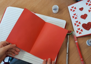 Dzieci wykonują kartki walentynkowe przy użyciu różnych materiałów: kolorowych serduszek, kartonów, taśmy ozdobnej.