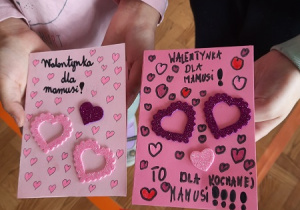 Dzieci prezentują wykonane przez siebie kartki walentynkowe. Na różowym kartonie ponaklejane kolorowe serduszka.