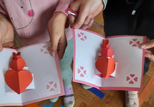 Dzieci prezentują wykonane przez siebie kartki walentynkowe. Na czerwony karton naklejona biała kartka ksero z powycinanymi małymi serduszkami i ponaklejanymi czerwonymi sercami.
