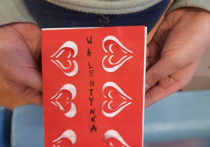 Dzieci prezentują wykonane przez siebie kartki walentynkowe. Czerwona kartka ksero z powycinanymi serduszkami naklejona na biały karton.
