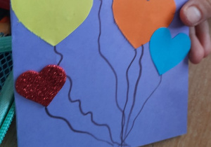 Dzieci prezentują wykonane przez siebie kartki walentynkowe. Na kolorowym kartonie ponaklejane kolorowe serduszka imitujące baloniki.