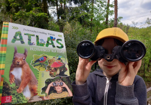 Zdjęcie przedstawia Kostka Roberta z kl. 2 b, który przez lornetkę podziwia osobliwości przyrody. W tle widać książkę „Atlas przyrodniczy dla dzieci”.
