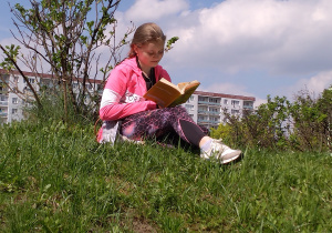 Wiktoria Filipczak z kl. 7 b czyta książkę siedząc na trawie. W tle widać bloki mieszkalne.