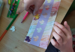 Dzieci przy użyciu kredek i kartonowych elementów wykonują zakładkę do książki o tematyce jesiennej.