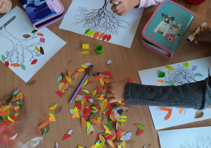Dzieci siedzą przy stolikach i wyklejają kontur drzewa kolorowymi papierkami w kształcie listków.