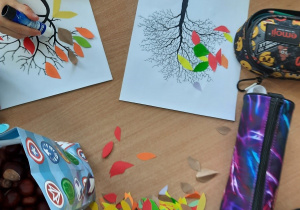 Dzieci siedzą przy stolikach i wyklejają kontur drzewa kolorowymi papierkami w kształcie listków.