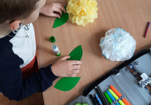 Chłopiec wykonuje kwiatki z białych i żółtych serwetek a listki z zielonego kartonu.