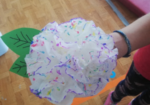 Dziewczynka prezentuje wykonany przez siebie kwiatek z białej bibuły, na którym namalowała kolorowe plamki, listki zrobiła z zielonego kartonu.
