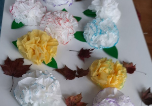 Prezentacja prac dzieci - Kwiatki wykonane z żółtych i białych serwetek na których namalowane są kolorowe obwódki, listki wykonane z zielonego kartonu.